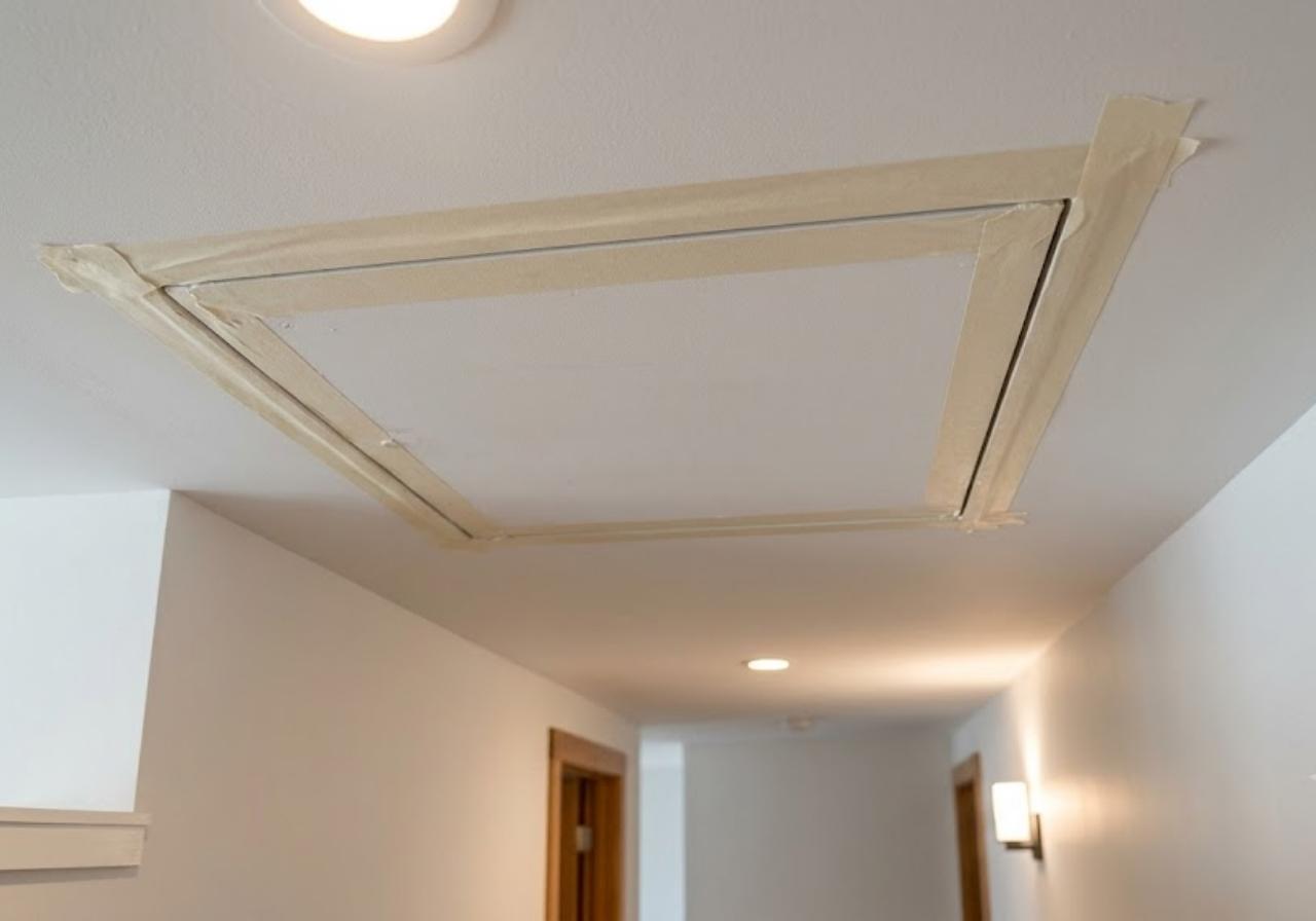 Rectangular ceiling attic access hatch outlined with masking tape in a hallway under recessed lighting.