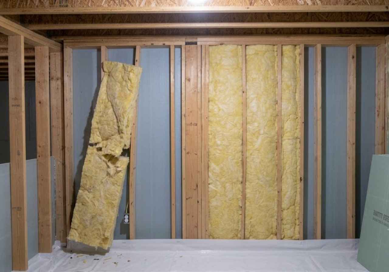 Unfinished basement wall with wooden studs and yellow fiberglass insulation between them, with a panel leaning against the studs.