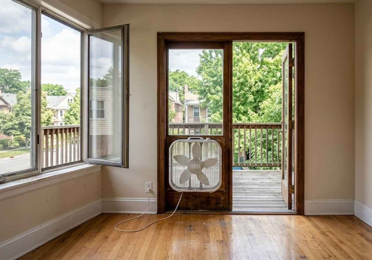 Empty beige room with hardwood floor, open door to a wooden balcony, and a portable fan perched in the doorway; large window to the left shows trees outside.